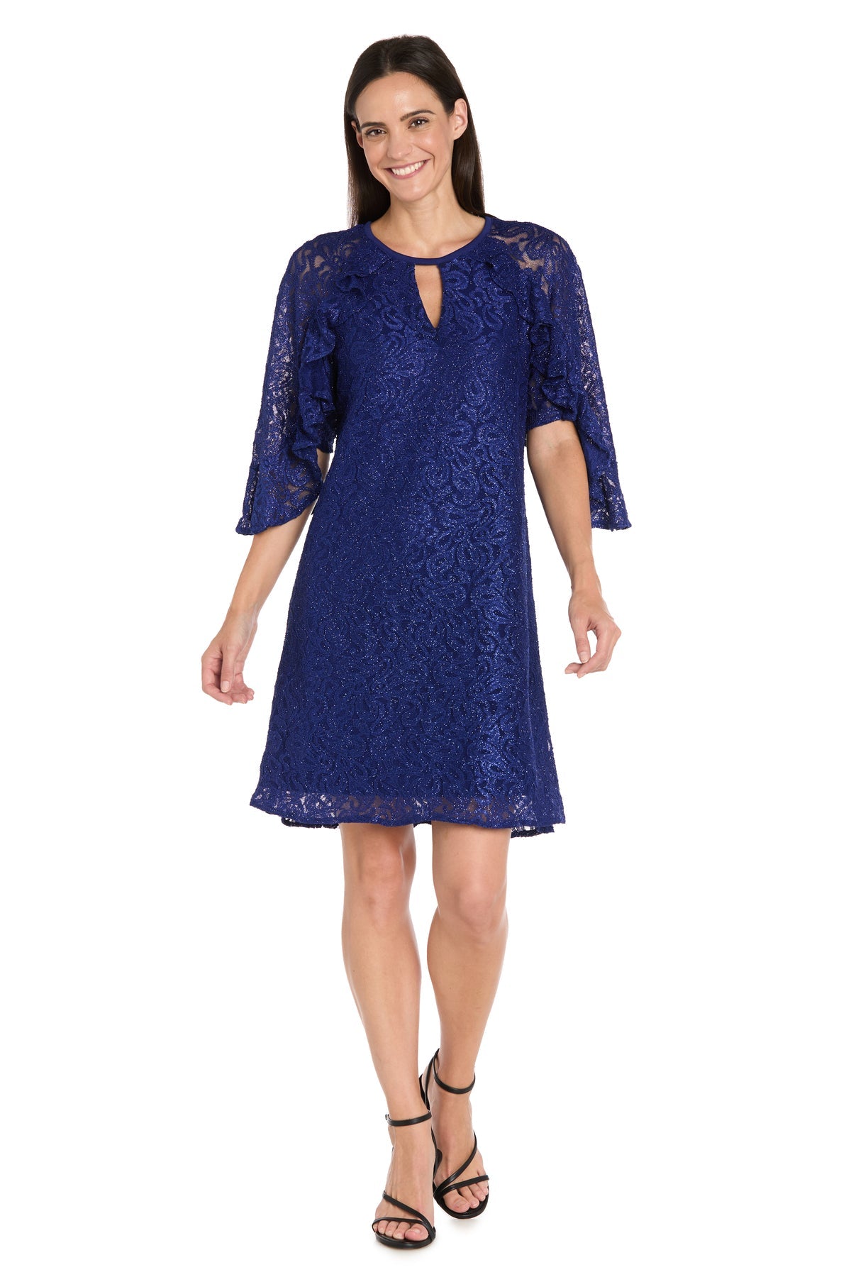 Woman models the R&M Richards 4226 Short Lace Semi-Formal Dress, featuring knee-length blue glitter lace, half sleeves, and paired with black strappy heels, smiling against a plain white background.